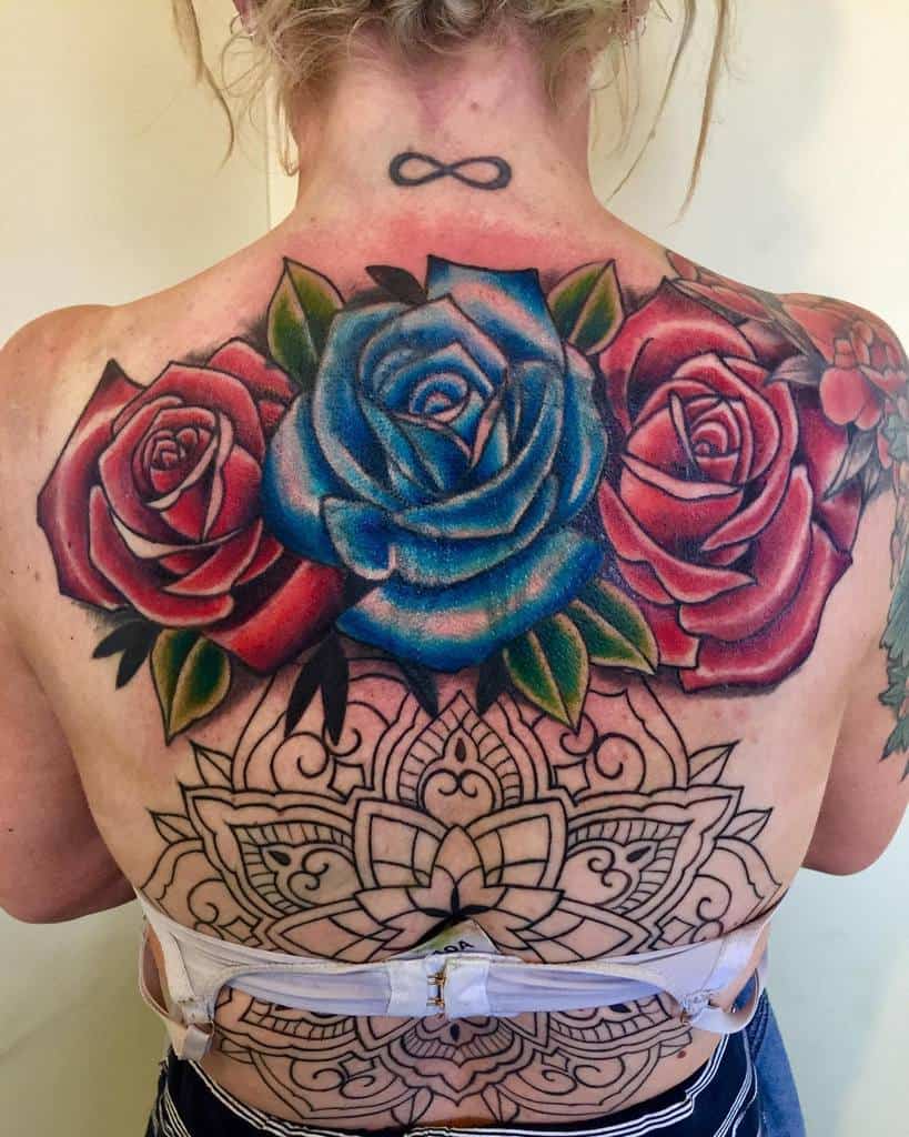 A bold and colorful tattoo on the back featuring a combination of red and blue roses, with vibrant shading and a mandala design beneath them, creating a striking and symmetrical composition