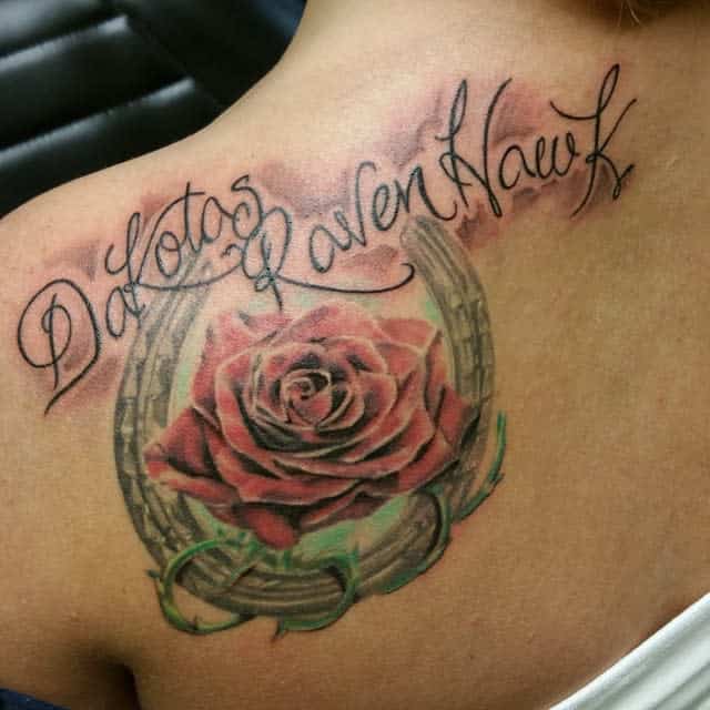 Back tattoo featuring a vibrant red rose within a horseshoe, adorned with green vines and flowing script text for a personalized touch