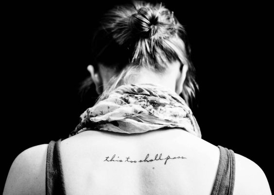 "This too shall pass" tattoo in cursive on the upper back, with the person wearing a scarf.
