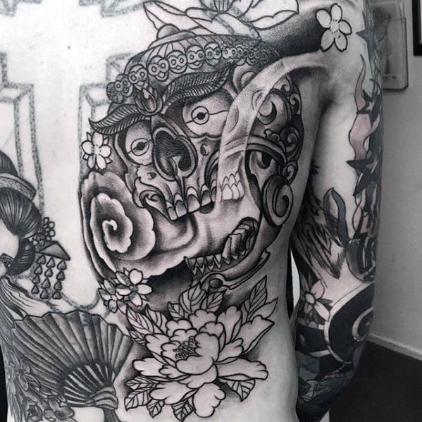 Black and grey Tibetan skull tattoo with floral elements and ornate headpiece, covering the upper back and shoulder.