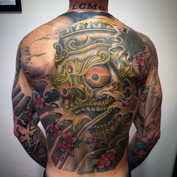 Colorful Tibetan skull tattoo with golden tones, swirling smoke, and pink cherry blossoms covering the full back and arms.