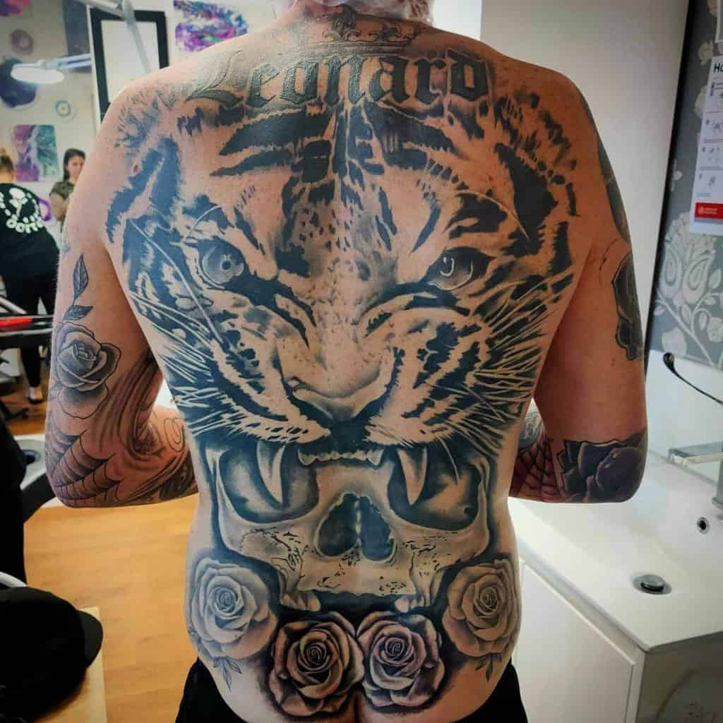 A stunning back tattoo artfully blends the fierce face of a tiger with the haunting presence of a skull, intertwined with delicate roses