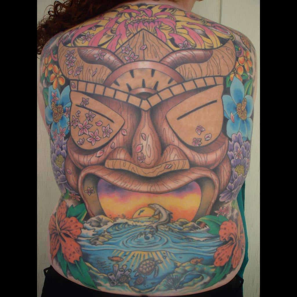 Vibrantly colored full-back Tiki tattoo, featuring a large mask surrounded by tropical flowers, a sunset scene with waves, and aquatic animals with reefs