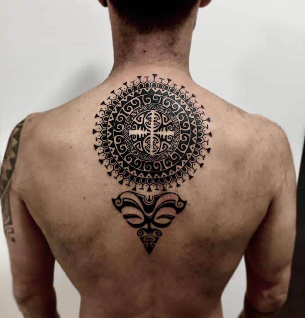 Bold black tribal tattoo, featuring a circular design with intricate patterns, surrounded by swirls, with a Tiki mask below, forming a triangular shape