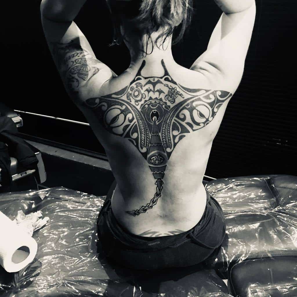 Black and grey Tiki tattoo, featuring a symmetrical tribal design with ornate patterns and a sharp, elongated tail-like element at the base