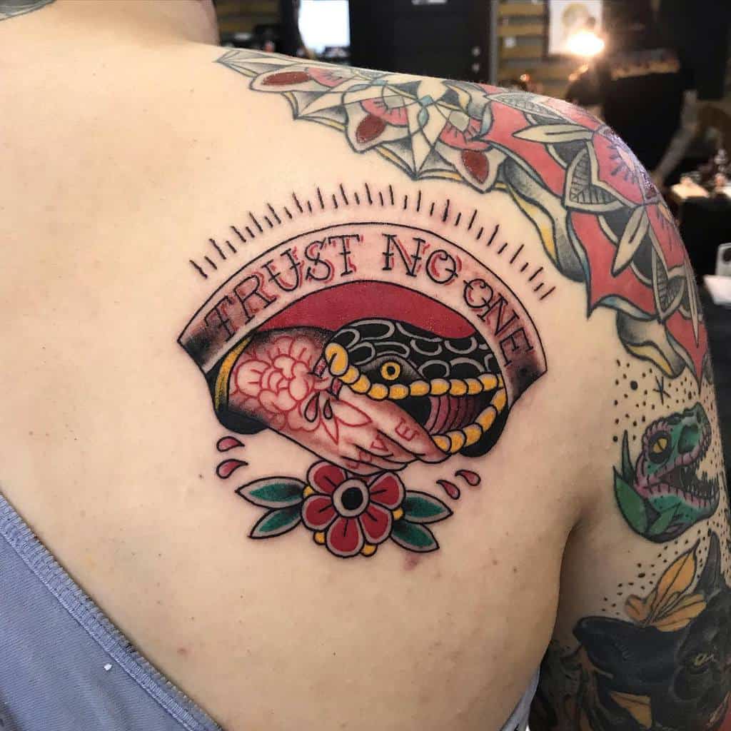 Bold upper-back tattoo featuring a snake handshake framed by the words 'Trust No One,' detailed with flowers and vibrant colors