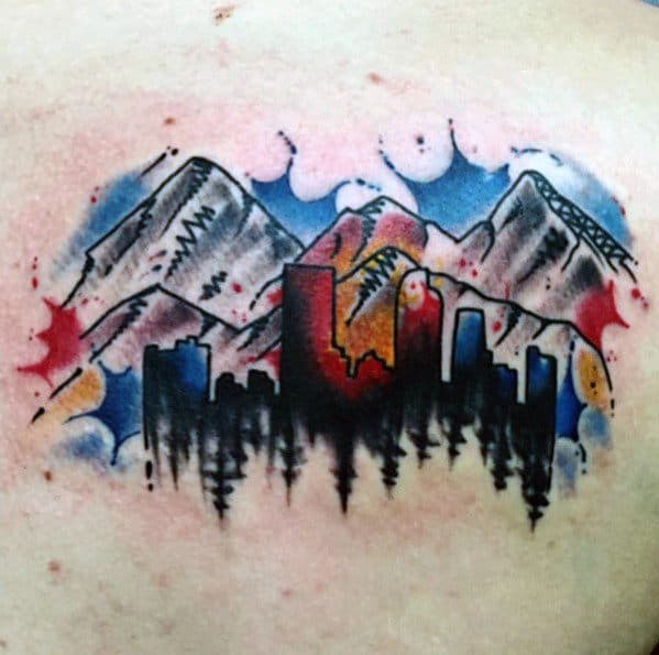 Watercolor-style Denver skyline tattoo with mountain backdrop and vibrant splashes of red, blue, and yellow behind the silhouette.