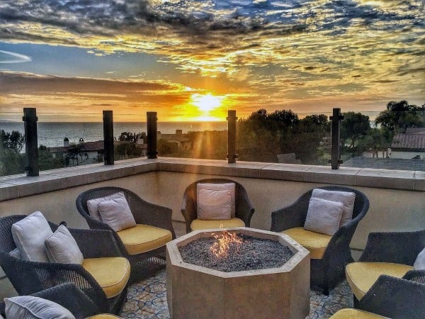 Wicker chairs encircle a cozy fire pit, offering views of a vibrant sunset sky and ocean