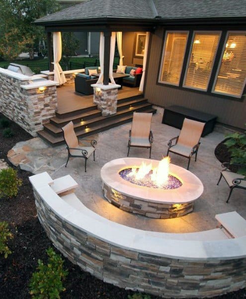 Outdoor patio with a fire pit, surrounded by chairs, near a raised deck with canopy seating
