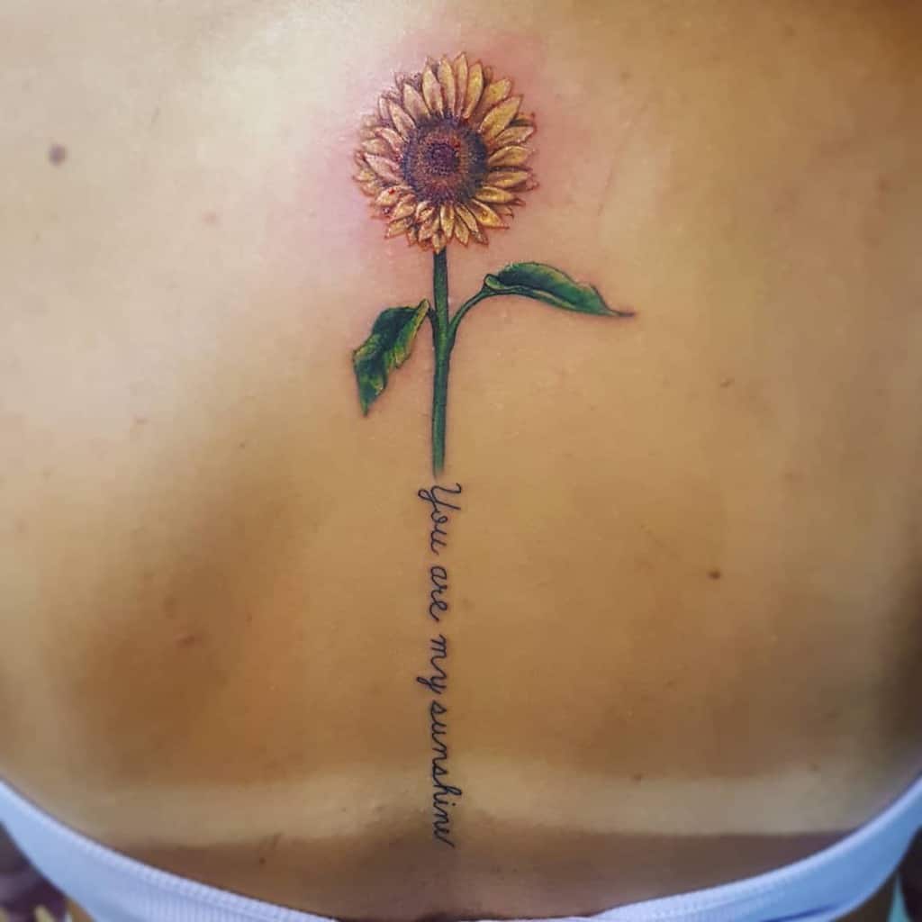 A vibrant sunflower tattoo on a person's back, featuring elegant lettering of the phrase "You are my sunshine" intertwined with the stem and petals to create a harmonious and meaningful design