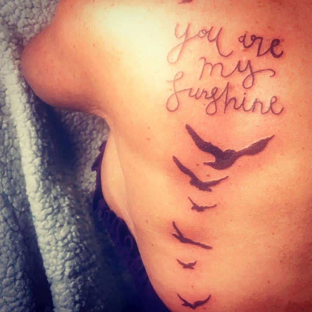 A tattoo design featuring the phrase "you are my sunshine" alongside flying birds on an upper back