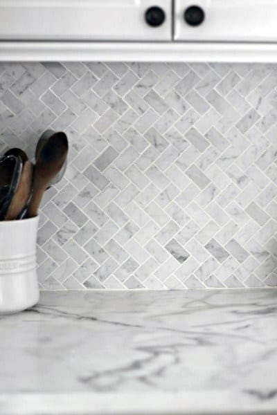Close-up of marble herringbone tile backsplash with marble countertop and utensil holder.