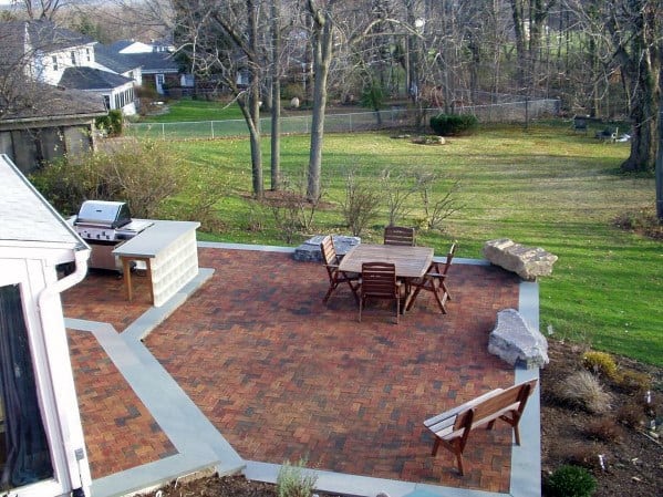 Charming brick patio with wooden furniture, grill, and large rocks, embraced by trees and grass