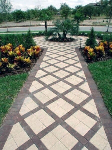 Geometric brick walkway with a checkerboard pattern, surrounded by lush greenery and colorful plants leading to a circular garden feature