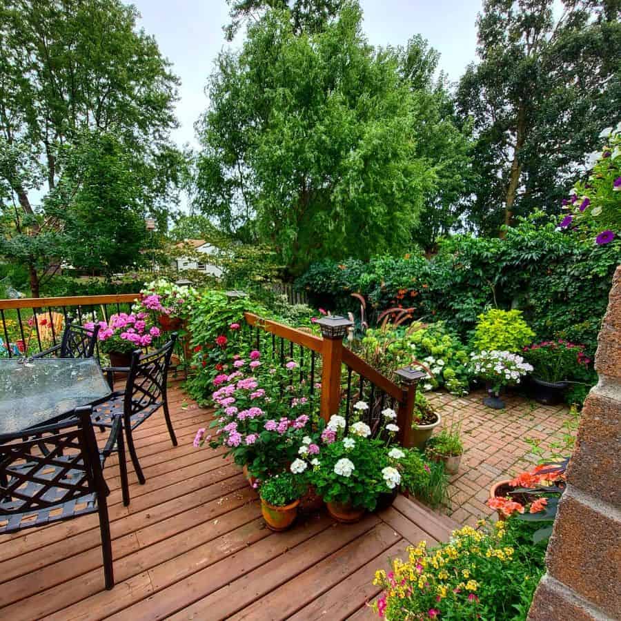 A wooden deck with a dining set, surrounded by lush green trees and colorful potted flowers in a garden setting