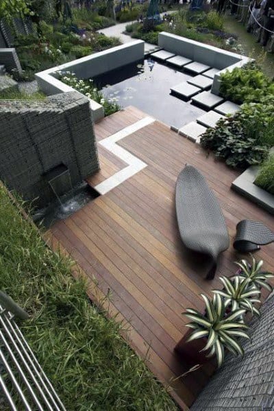 Wooden deck with modern seating, water feature, and lush garden surrounding a pond.