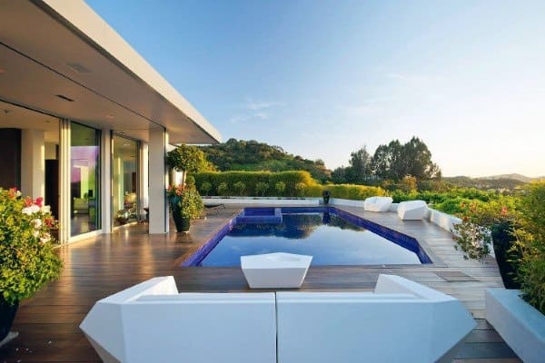 Modern wooden pool deck with white seating, lush greenery, and scenic hillside view.