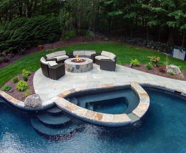 Circular patio with seating and a fire pit beside a pool, surrounded by greenery