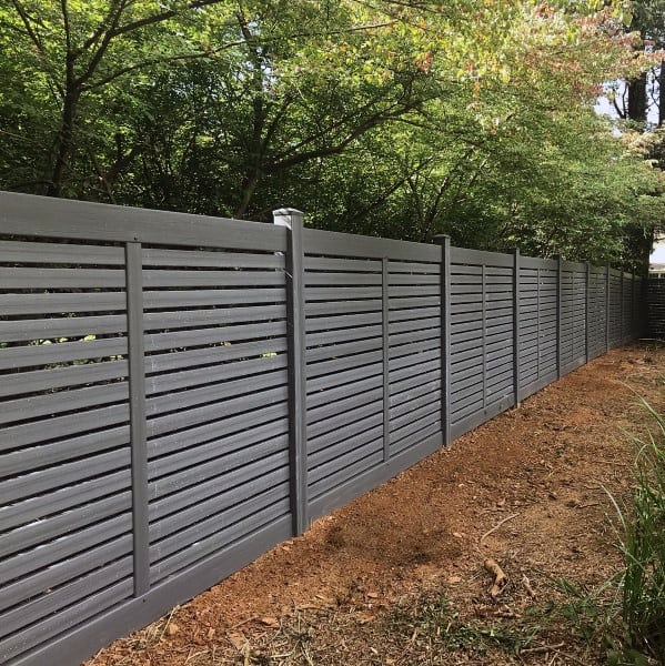 Modern grey vinyl backyard fence with horizontal slats, surrounded by trees and dirt path.