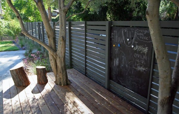 Modern metal backyard fence with a built-in chalkboard and wooden deck seating area.