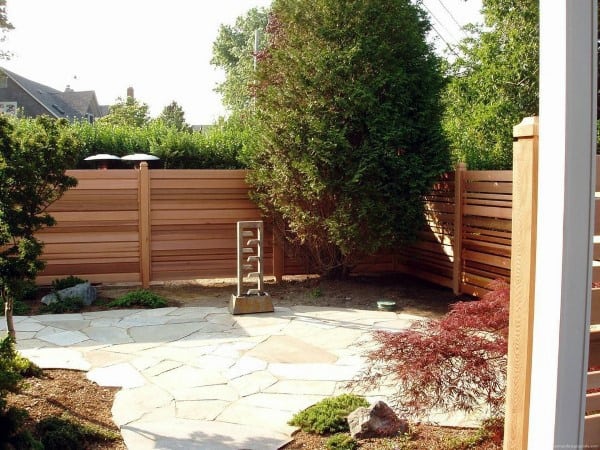 Horizontal wooden fence surrounding a small backyard with a stone patio and lush greenery.