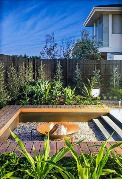 Modern backyard with a fire pit, wooden seating, lush greenery, and a house in the background at dusk
