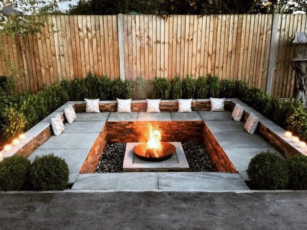 Cozy fire pit area with stone seating, cushions, surrounded by a wooden fence and lush greenery