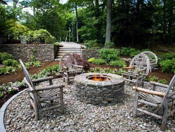 Rustic stone fire pit surrounded by wooden chairs in a garden with lush greenery and a stone wall