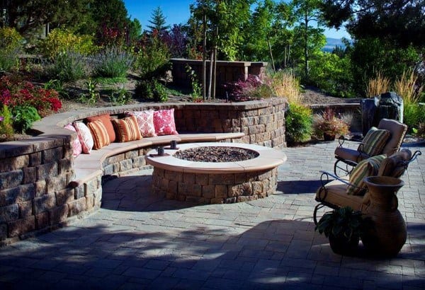 Cozy outdoor patio with a circular fire pit, curved stone bench, colorful cushions, and lush surrounding greenery