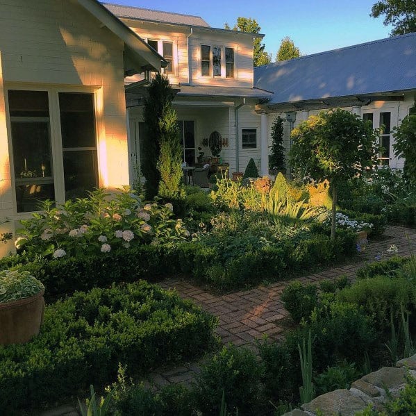 Brick walkway weaving through a lush garden with neatly trimmed bushes, vibrant flowers, and a cozy home in the background