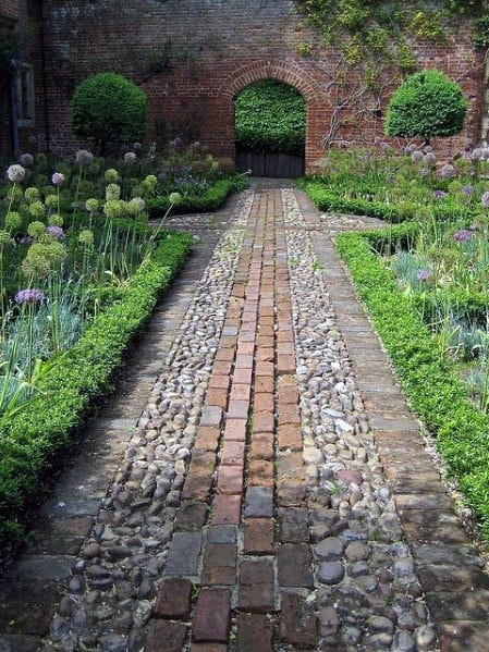 A brick and pebble walkway winds through a garden with an archway and greenery