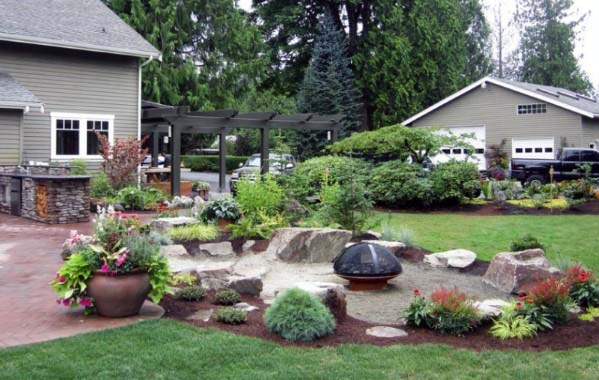 Backyard with a garden, fire pit, and pergola, adjacent to a house and garage surrounded by lush greenery