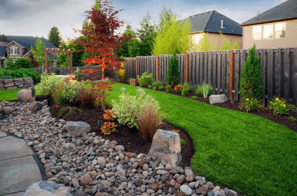 Beautiful backyard with a curved river rock border, vibrant plants, and a well-maintained lawn, surrounded by a wooden fence