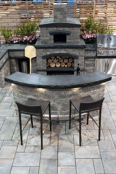 Rustic outdoor stone bar with a wood-burning oven and wicker chairs on a paved patio.
