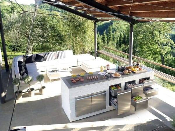 Outdoor kitchen with a white island, stainless steel appliances, and lounge area set in a lush outdoor landscape