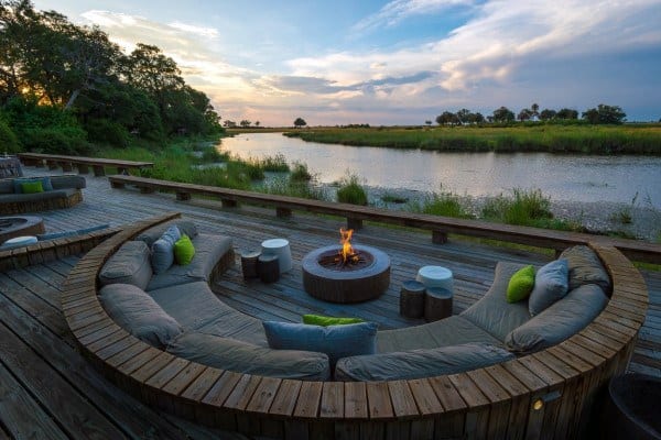 Wooden deck with circular seating, firepit, and scenic waterfront view at sunset.