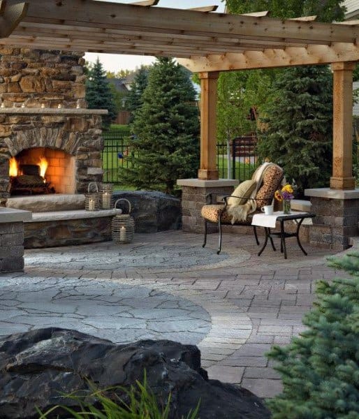 Cozy patio with a lit stone fireplace, chair, flower-topped table, and trees in the background