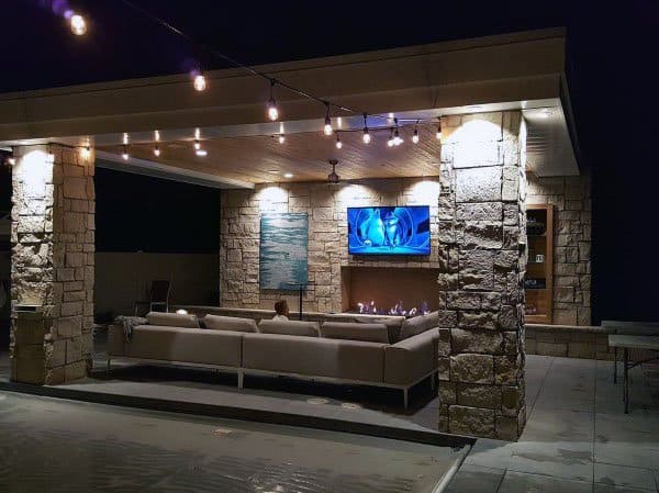 Modern stone pavilion with cozy seating, outdoor fireplace, and a mounted TV, illuminated at night.