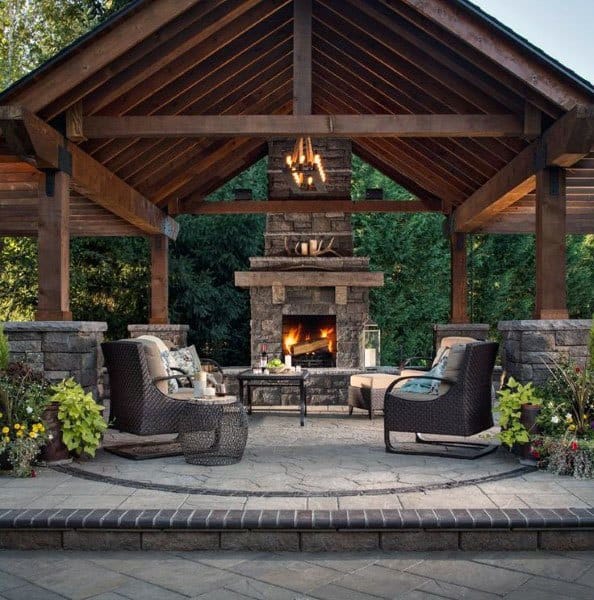 Stone pavilion with a cozy fireplace, wicker seating, and chandelier lighting in a lush garden setting.