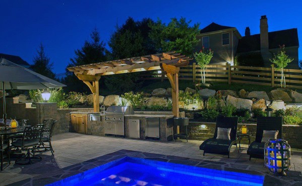 Nighttime backyard scene with a pergola-covered outdoor kitchen, built-in grill, patio furniture, and illuminated pool