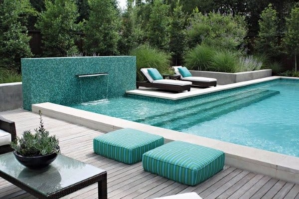 Modern pool with a sleek wall waterfall feature, lounge chairs, and lush greenery.