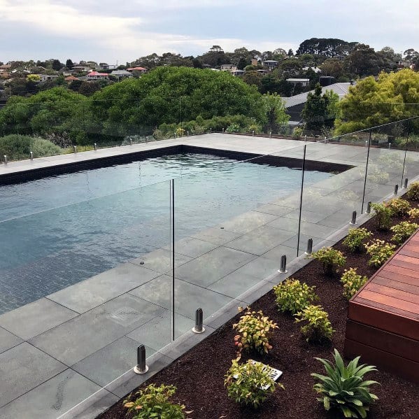 Modern outdoor pool with glass fencing, surrounded by greenery and a view of houses in the background
