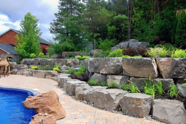 Backyard pool area with a sloped rock wall, layered greenery, and a paved patio creating a natural and relaxing outdoor retreat