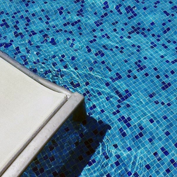 Close-up of blue mosaic pool tiles with a white lounge chair on the water’s edge.