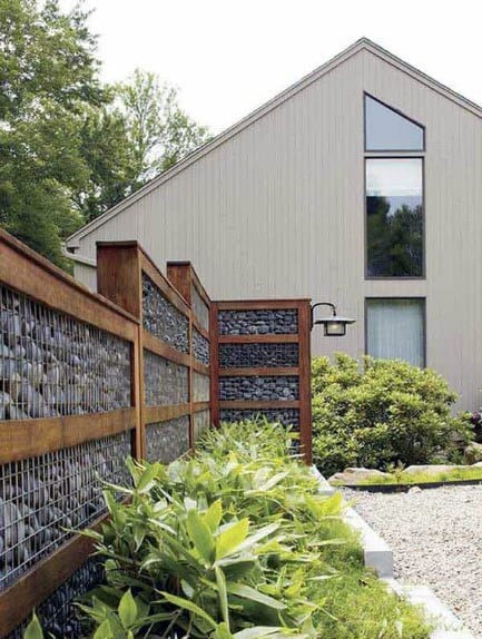 Gabion fence with wooden frames filled with stones, bordering a modern home with a gravel pathway.