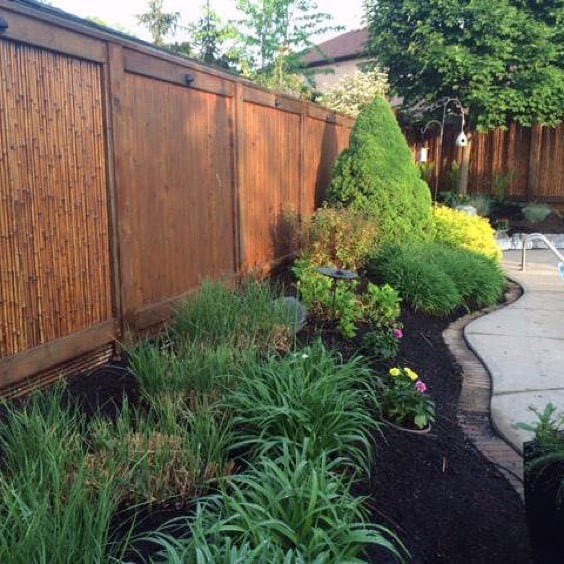 Bamboo privacy fence with wooden frame in a landscaped backyard featuring greenery and flower beds.