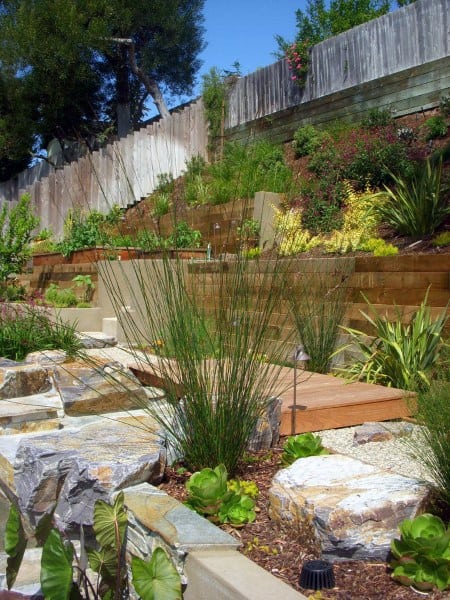 Tiered wooden and stone retaining walls with vibrant plants and a small wooden pathway.