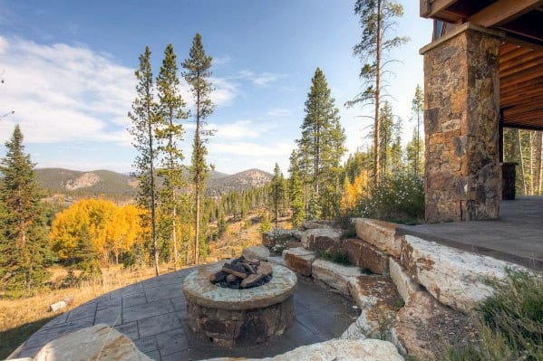 Rustic backyard patio on a slope with a stone fire pit, natural rock seating, and stunning mountain views surrounded by tall pines