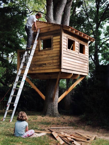 Person on ladder building treehouse; child sits on ground watching
