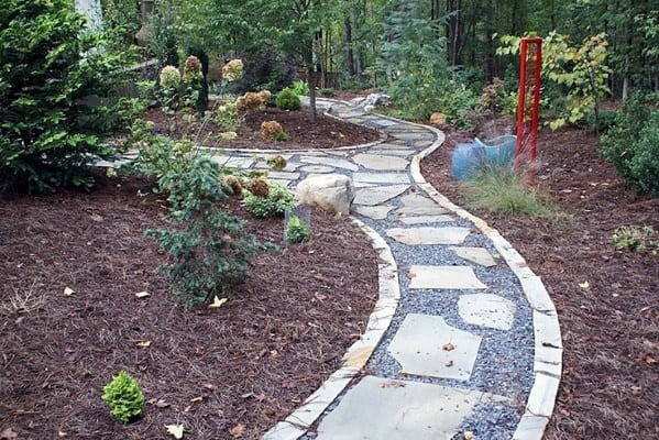 A winding stone pathway with gravel edging, surrounded by lush greenery and mulch in a peaceful garden setting